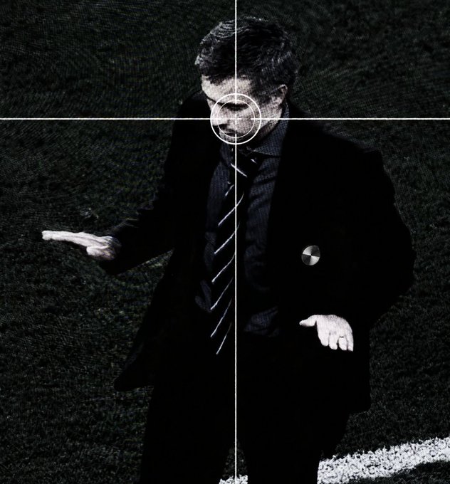 A man in a dark suit and striped tie stands on grass with his hands outstretched, overlaid with a sniper-style crosshair targeting his head and a circular marker on his jacket pocket.
