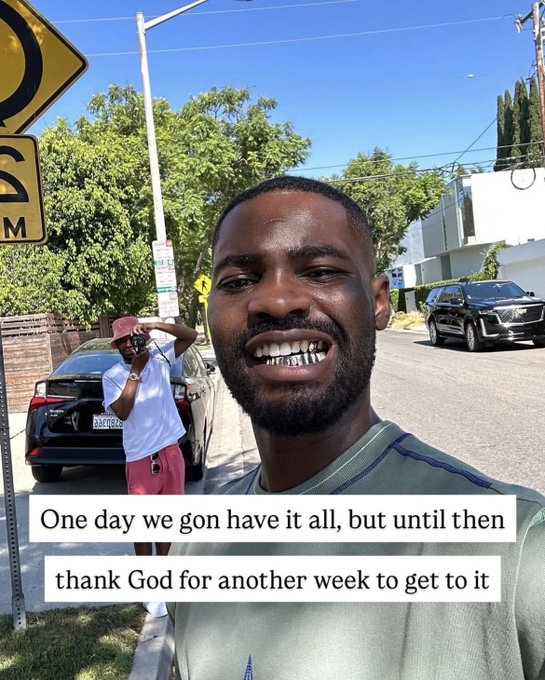 A man taking a selfie outdoors on a sunny day with a caption reading 'One day we gon have it all, but until then thank God for another week to get to it.'
