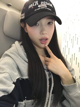 A young woman wearing a black cap and gray jacket takes a selfie while making a peace sign with her finger.