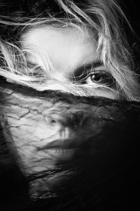 A black and white close-up portrait of a person's face partially covered by a sheer veil or fabric, with light hair blowing across the frame and one visible eye.