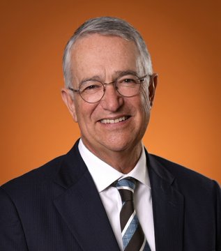 The image features an older man wearing glasses and a suit jacket with a tie. He has a smile on his face, giving off a friendly vibe. The background of the photo is orange, which contrasts nicely with the man's attire. There are no texts or other objects in the image, making it a simple yet visually appealing composition that highlights the subject matter.