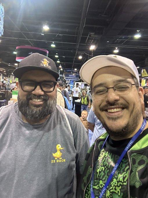 Two smiling men pose for a selfie at what appears to be a convention; the man on the left wears a cap and a 'ZZ Duck' t-shirt, while the man on the right wears glasses and a green hoodie with a 'ByteFed' lanyard.