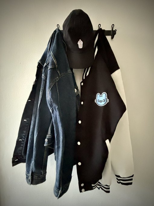 A black cap, denim jacket, and a black-and-white varsity jacket with a cartoon mouse design hanging on a wall coat rack.
