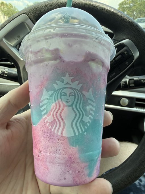 A hand holds up a Starbucks "Unicorn Frappuccino" inside a car, showing its swirled pink and blue blended contents through the clear cup featuring the iconic Starbucks siren logo. The caption's critique of the drink being "gross af" directly reflects the muddy, unappetizing appearance of the mixed colorful beverage once blended together.