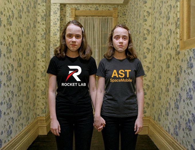 Two twin girls stand side by side in a hallway with floral wallpaper, wearing shirts branded with "Rocket Lab" and "AST SpaceMobile" logos.