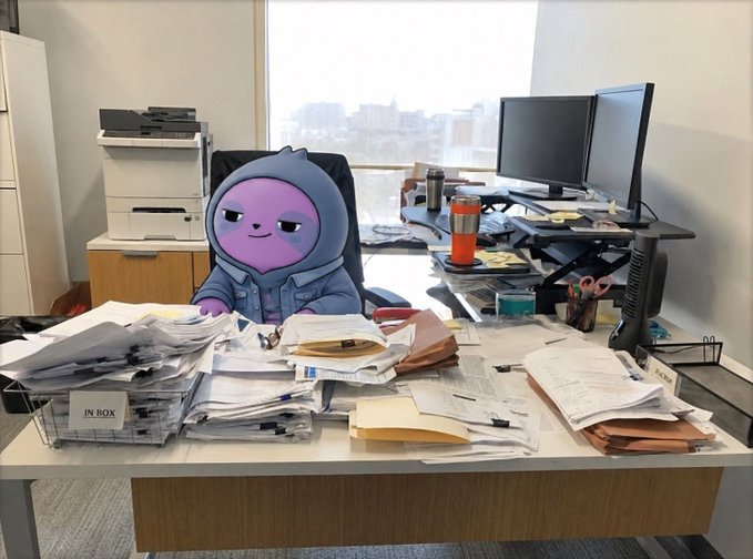 Office scene with a purple cartoon character in a hoodie sitting at a cluttered desk overflowing with papers, next to a wire basket labeled 'IN BOX' and dual monitors.