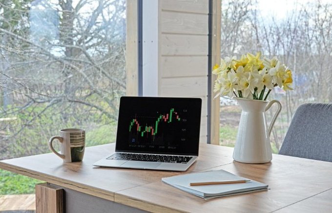 A wooden desk by a window overlooking a forest, with a laptop displaying a candlestick chart, a coffee mug, a vase of yellow daffodils, and a notebook with a pencil.