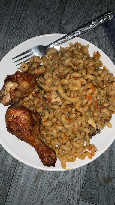 A white bowl containing elbow macaroni pasta with what appears to be vegetables, served alongside two pieces of fried chicken on the bone, with a fork resting on the plate.