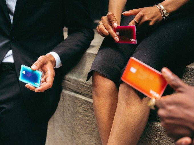 Three people in business attire hold colorful Visa credit cards, including blue, red, and orange ones, while seated on stone steps.