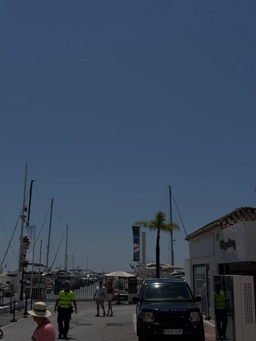 A sunny marina scene shows a black Land Rover (license plate 7115 GJN) parked near a building labeled "William," with sailboat masts, a palm tree, and people strolling under a clear blue sky—capturing the leisurely, wealth-adjacent lifestyle vibe that complements the "Gm (get money)" post.