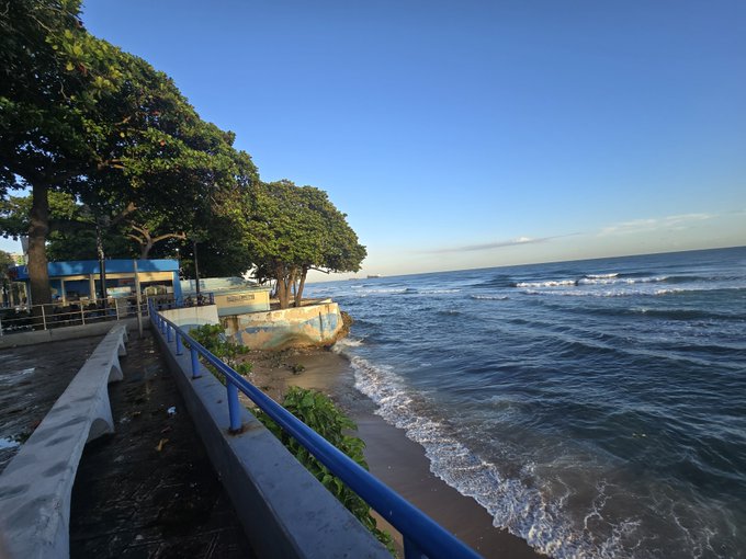 Coastal scene at a paved seaside promenade with blue railings, trees, and waves rolling onto the beach under a bright blue sky.