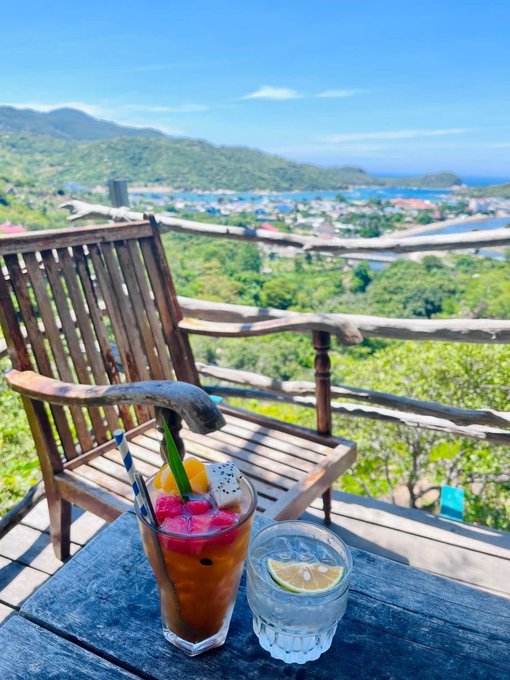 Scenic view from a wooden deck featuring a fruit cocktail and a glass of water with lime, overlooking a tropical coastal town with green hills and blue ocean in the background.