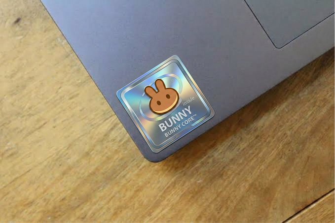 A holographic "Bunny Core™" sticker adorns the corner of a gray laptop resting on a wooden surface, playfully suggesting the user's computer harbors this "oldest flex" as Mystery Stock #15 from pancakeswap.finance/stocks.