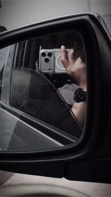A close-up of a car's side mirror reflecting a hand holding a smartphone with a triple-camera setup, along with an Apple Watch on the wrist.