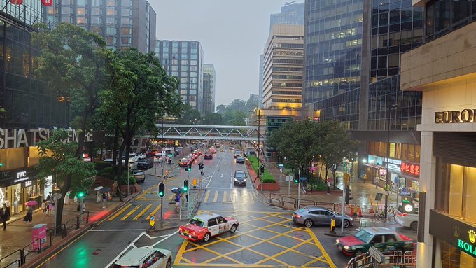 A rainy urban street scene in Hong Kong with heavy traffic, pedestrians carrying umbrellas, and visible storefronts including IWC and Euro, capturing the wet weather conditions mentioned in the post.