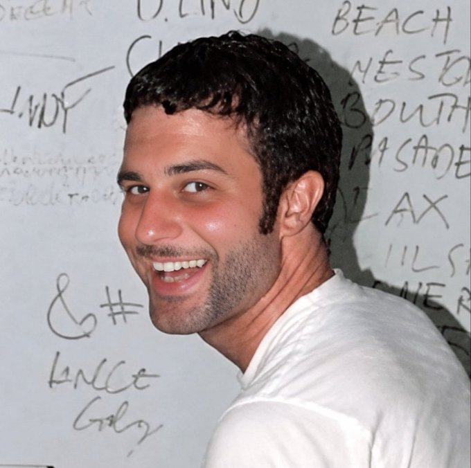 A smiling young man with dark curly hair and stubble stands in front of a white wall covered in handwritten graffiti and messages.