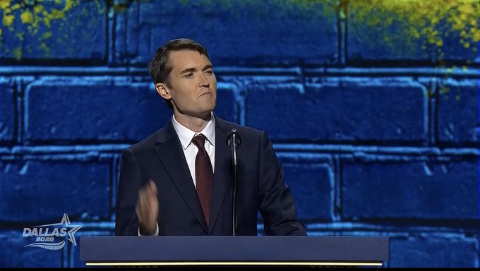A man in a dark suit and tie standing at a podium speaking into a microphone, with a blue brick wall backdrop and 'DALLAS COWBOYS' logo visible in the bottom left corner.