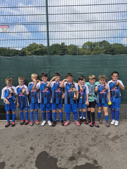 A youth soccer team in blue uniforms posing together with trophies and medals, standing in front of a green fence with a 'DANGER Do not climb' sign.