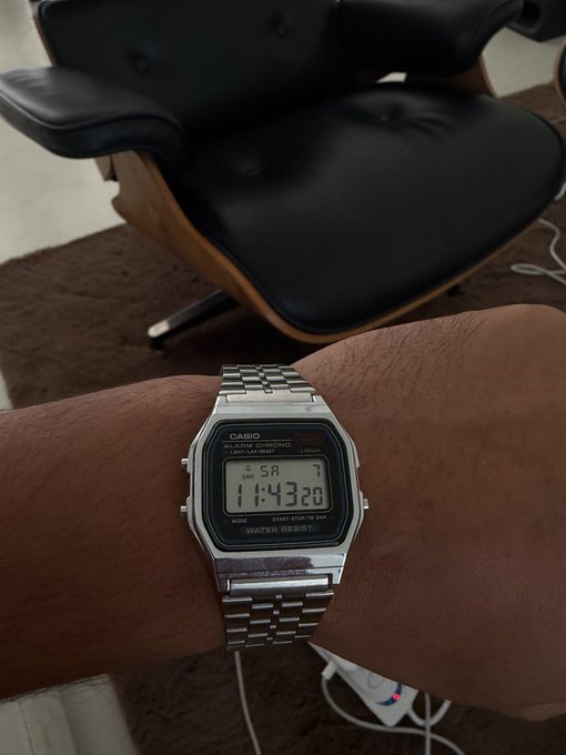 A wrist flexes a silver Casio digital watch displaying "SA 11:43 20" in front of a black leather mid-century modern chair, showcasing the classic timepiece as the main flex.