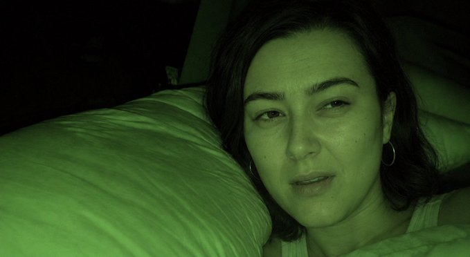 A green-tinted night vision photo of a woman with dark hair lying on a pillow, looking directly at the camera.