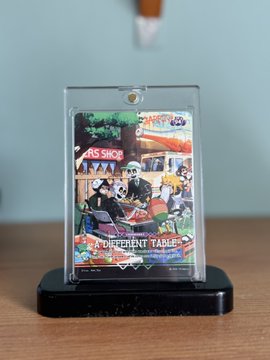 A clear acrylic display case on a black stand showcases a colorful trading card with anime-style characters, featuring visible text like 'THE COFFEE SHOP' and 'The Different Table'.