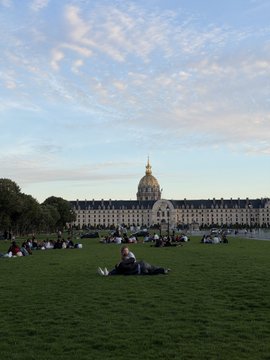 A scenic view of a large grassy field with people relaxing on it, with the golden dome of Les Invalides in Paris visible in the background under a cloudy sky.
