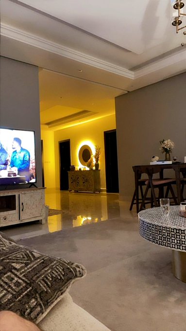 A dimly lit living room interior with a TV on the left, a yellow accent wall with a round mirror, a side table, and dining chairs visible in the background.
