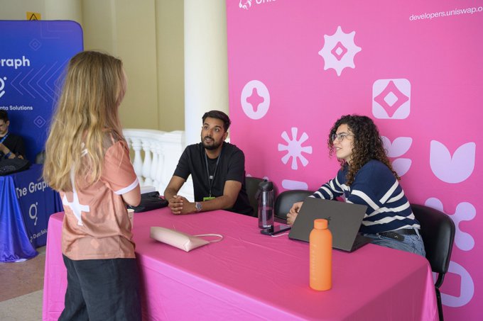 A networking event at what appears to be a Uniswap developers conference, where two people sit at a pink table with laptops while a woman in a pink and white top stands talking to them.