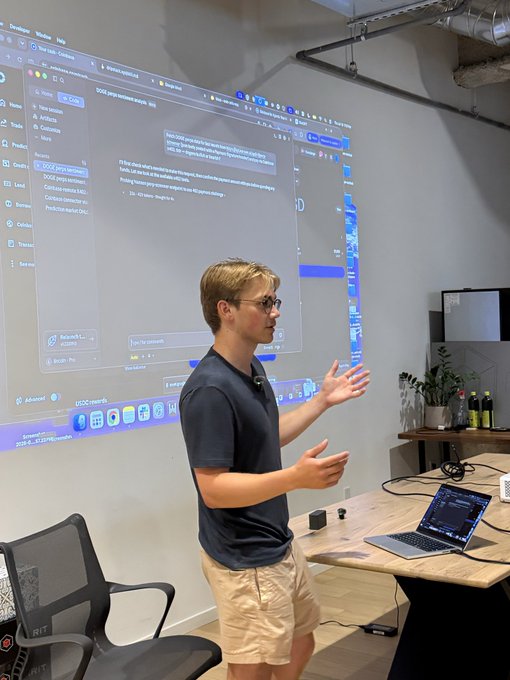 A man in glasses and a dark t-shirt presents in an office setting, standing before a large projected screen displaying a Notion workspace interface, with a MacBook on the wooden desk nearby.