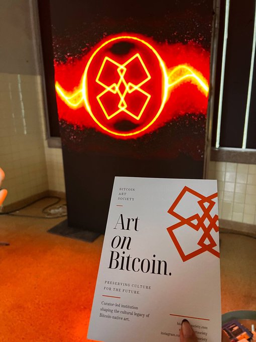 A hand holds a brochure titled "Art on Bitcoin. Preserving Culture for the Future" by the Bitcoin Art Society, displayed in front of a glowing red and yellow art piece featuring geometric symbols—visually echoing the post's message about lineage being permanently inscribed on the Bitcoin blockchain.