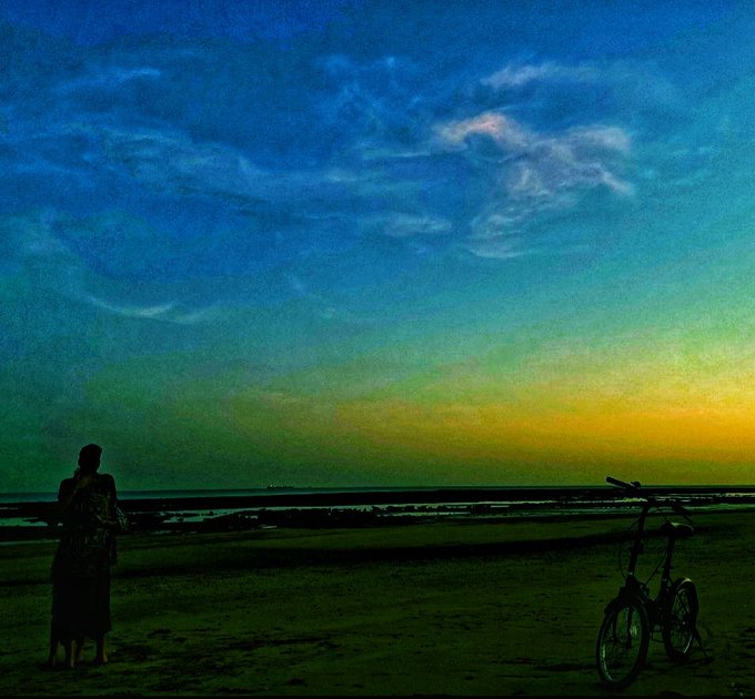 A silhouetted scene of a person standing on a beach next to a bicycle, watching a vibrant sunset over the ocean.