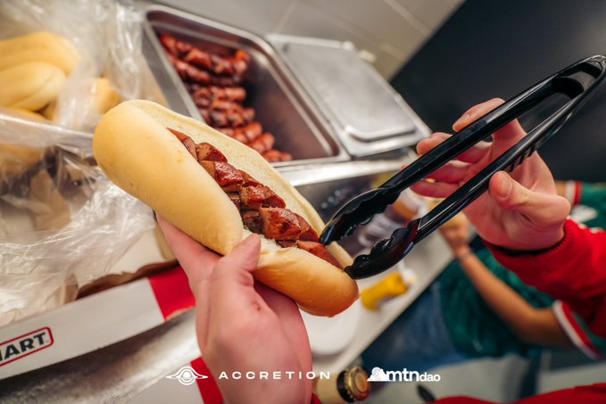 Two hands assemble a hot dog topped with bacon bits using black tongs, with metal serving trays of additional toppings and buns visible in the background. The image is watermarked with "ACCRETION" and "mtndao" logos, supporting the post's playful claim about Accretion's hot dogs being the "Highest ROI on Solana."