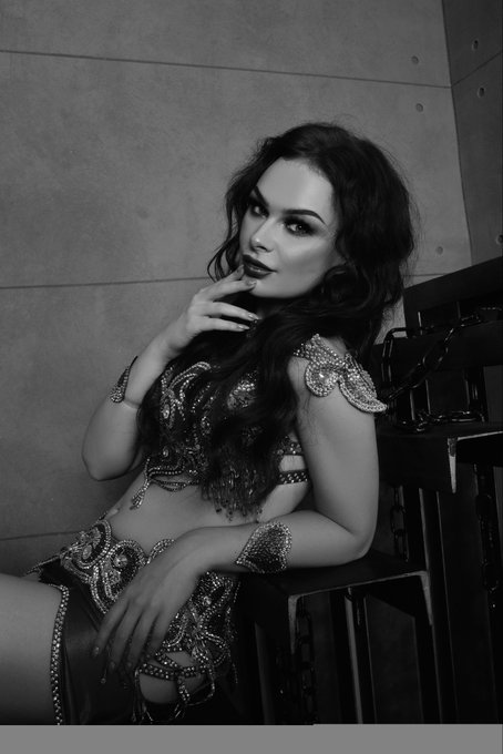 A black-and-white portrait shows a woman with long wavy hair in ornate embellished clothing, posing with one hand near her face.