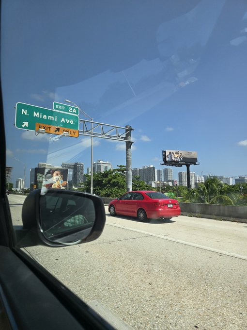 A view from inside a car showing highway exit signs for Exit 2A N. Miami Ave., with a red sedan driving on the road and a city skyline in the background.