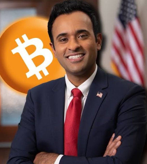 AI-generated or stylized image of a smiling South Asian man in a navy suit and red tie with an American flag pin, posed in front of a Bitcoin logo and U.S. flag backdrop.