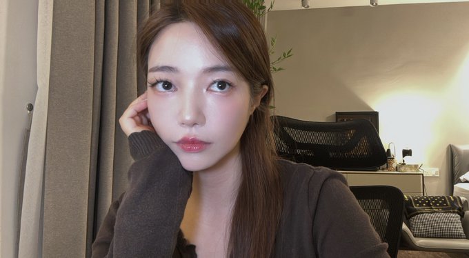 A woman with long brown hair and makeup poses for a selfie in a room with a desk, chair, and beige curtains in the background.