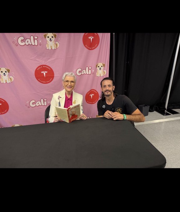 An older woman in a white jacket and a younger man in a dark t-shirt sit at a black table in front of a pink backdrop with 'Cali' and 'Silicon Valley' logos, with the woman holding an open book.
