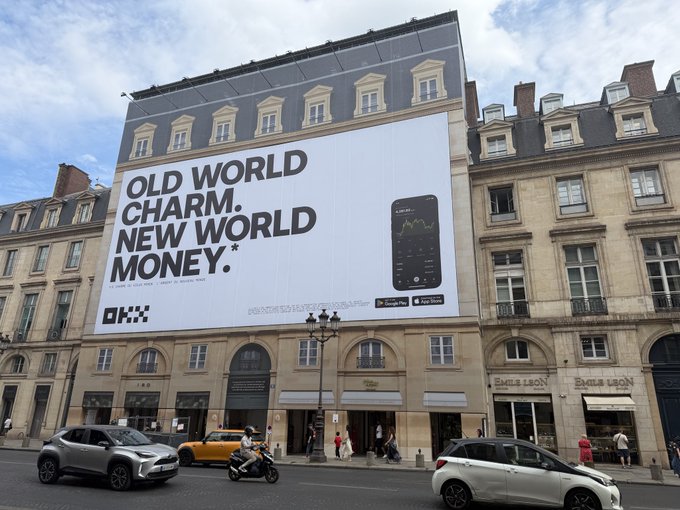 A large building billboard advertises OKX with the text 'OLD WORLD CHARM. NEW WORLD MONEY.*' alongside an image of a smartphone displaying a trading chart.