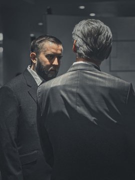 A cinematic, dimly lit scene showing two men in suits facing each other, with the camera positioned behind the gray-haired man looking at his bearded counterpart.