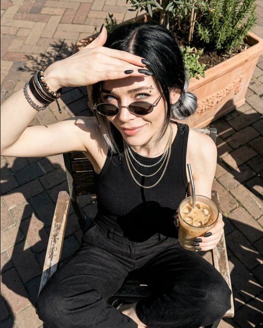 A person with black and white hair wearing sunglasses and a black tank top sits outdoors holding an iced coffee, posing with their hand shielding the sun.