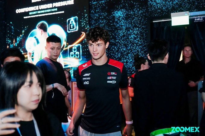A photograph of two young men, one wearing a Haas F1 team shirt, standing in front of a screen displaying "CONFIDENCE UNDER PRESSURE" and the Zoomex logo.