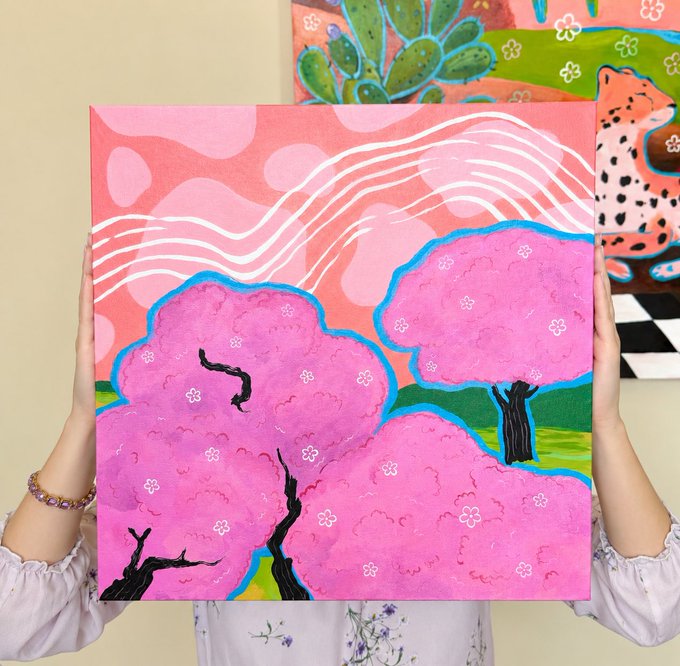 A pair of hands holding up a colorful acrylic painting of pink cherry blossom trees against a pink sky with white cloud lines, with another painting featuring a leopard and cactus visible in the background.