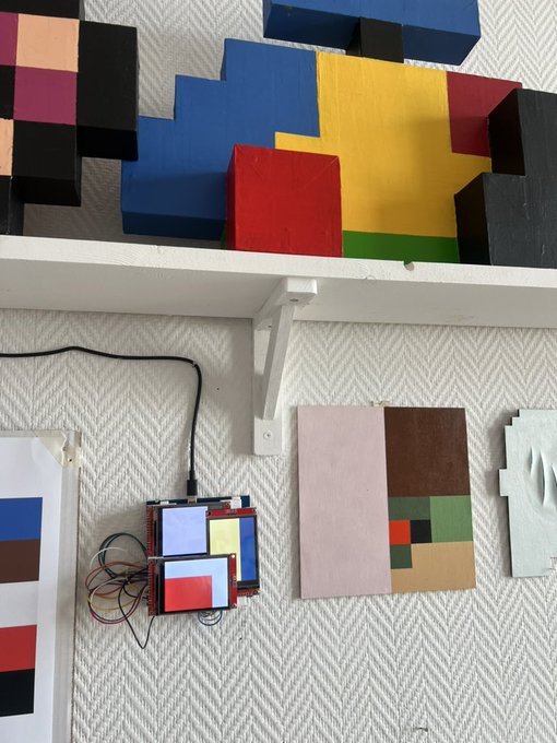 An art display with colorful geometric wooden blocks arranged on a white shelf above smaller pixel-art style paintings mounted on a textured white wall.