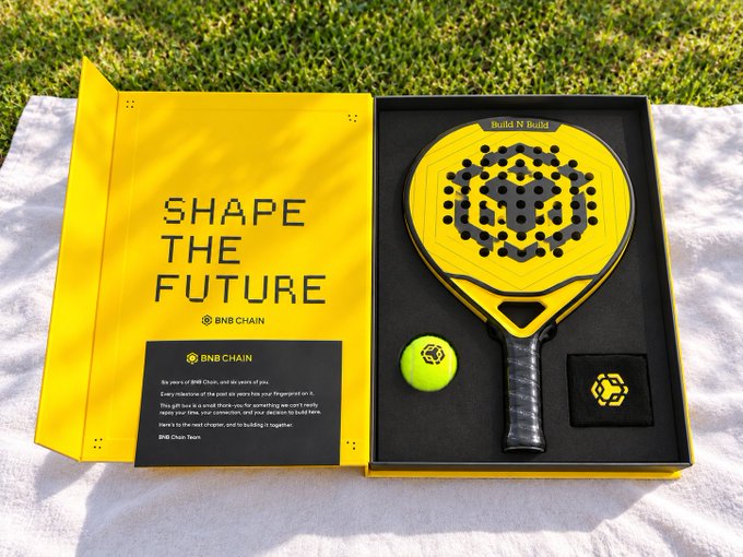 An opened yellow presentation box on a white towel over grass reveals a yellow and black 'Build N Build' padel racket, a yellow tennis ball, and a black wristband. The inside cover of the box reads 'SHAPE THE FUTURE' alongside the BNB CHAIN logo and a signed message.