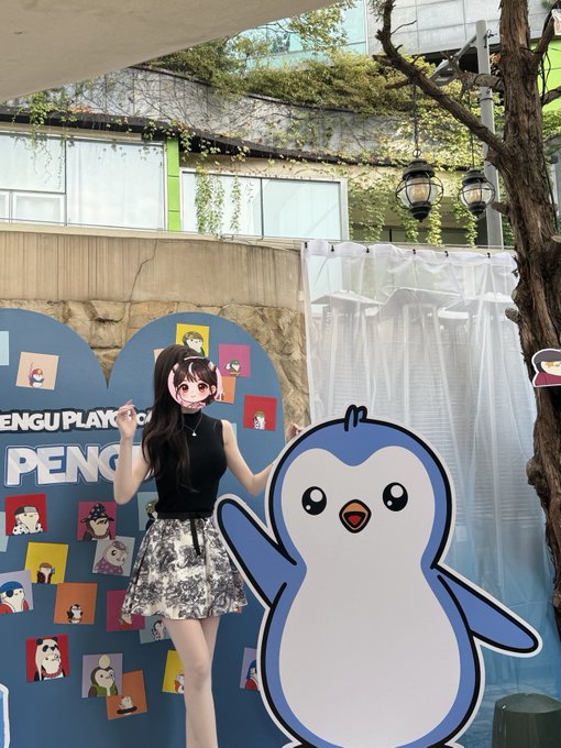 A young woman wearing a penguin face mask poses next to a large penguin cutout at what appears to be a 'PENGU Playground' event.