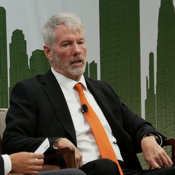 A man with a gray beard, wearing a dark suit and bright orange tie, sits on a stage in front of a green cityscape backdrop, appearing to be speaking at a panel. The image likely depicts Michael Saylor, the Strategy executive, as he discusses his vision for a bitcoin-backed stablecoin during an appearance at ParaFi Investor Day.