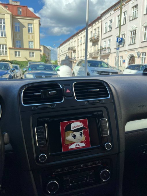 A car dashboard displays a RedStone sticker on its infotainment screen, humorously framed as the driver's trusted navigation oracle. Parked on a city street with European architecture in the background, the image visually reinforces the joke about treating the RedStone sticker as essential to avoiding getting lost.