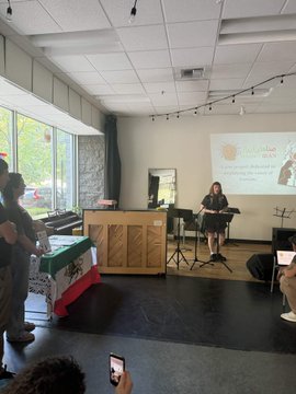 A musician plays a flute on a small stage in a bright room while a small audience watches, with a banner on a nearby table and a projector screen in the background.