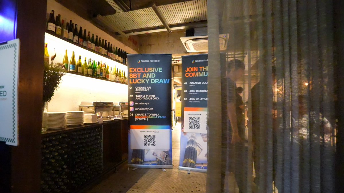 The image shows a group of people gathered around a table with various items such as pizza boxes, cards, and banners. The scene appears to be set at a restaurant or a social gathering where people are enjoying themselves. There are two large banners prominently displayed in the room, one on each side, possibly advertising an event or promotion related to the gathering. A man is seen holding a card, which could be part of a game or a special offer for the attendees. The atmosphere seems lively and engaging as everyone participates in the activities happening around them.