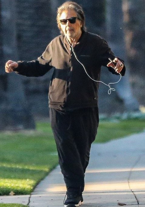 Actor Al Pacino walks along a sunlit sidewalk wearing sunglasses and dark clothing, with wired earphones in his ears and a smartphone in his hand.